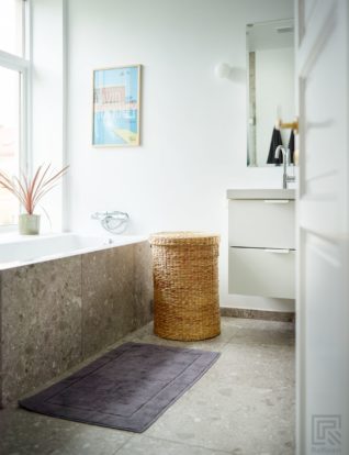 Innbygd badekar under vinduet med terrassoinspirerte fliser i front, som på gulvet. Den hvite innrednignen til vasken har to store skuffer. Hvit vegglampe ved siden av speilet på den hvitmalte veggen.