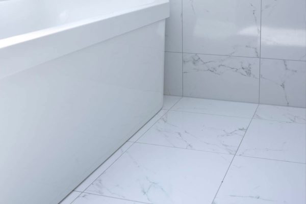 Frittstående hvitt badekar under skråtaket, med hylle innerst med marmorfliser, som på gulvet. På hyllen ligger det hvite brettede håndklær og står en såpeflaske.