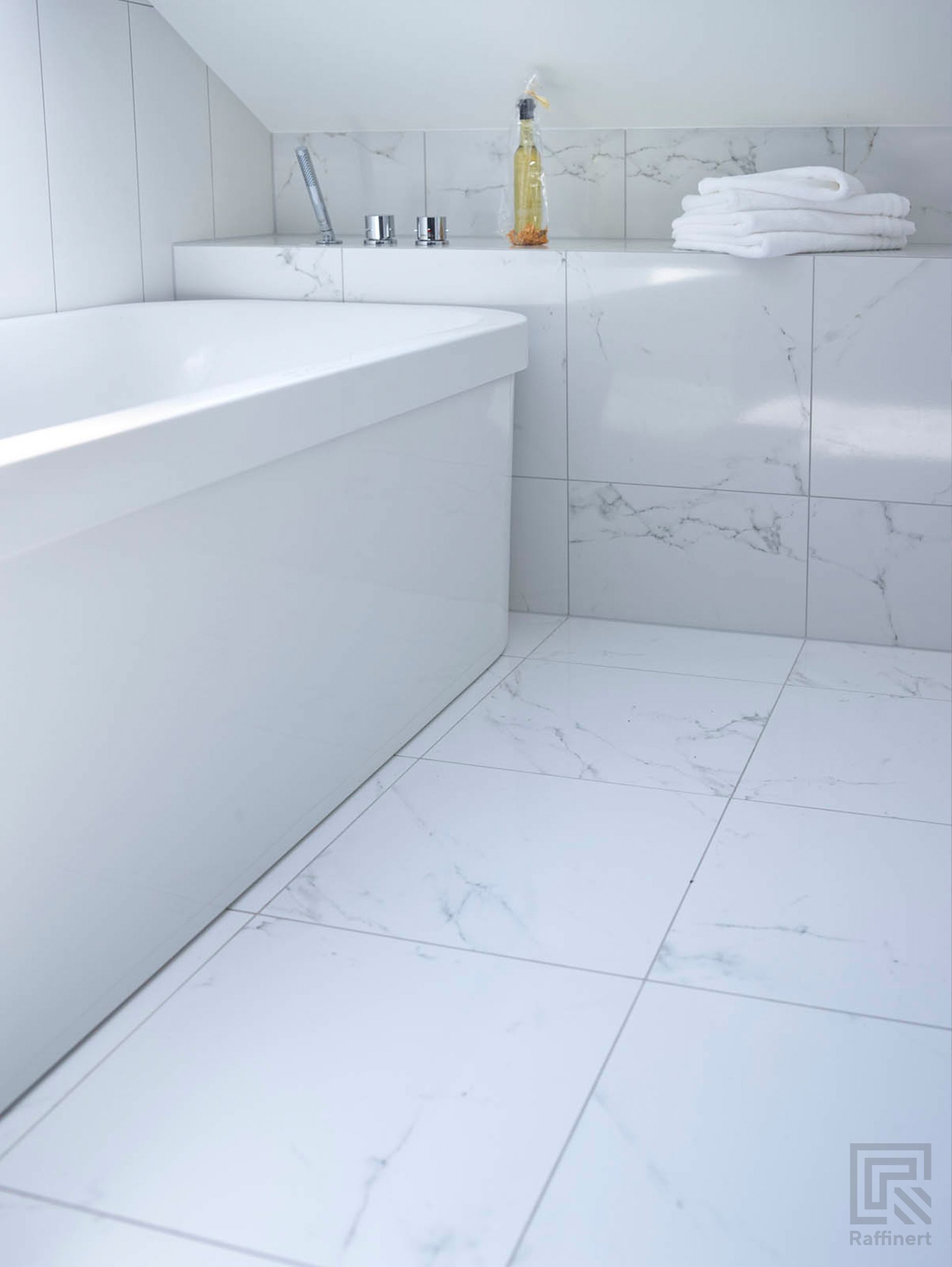 Frittstående hvitt badekar under skråtaket, med hylle innerst med marmorfliser, som på gulvet. På hyllen ligger det hvite brettede håndklær og står en såpeflaske.