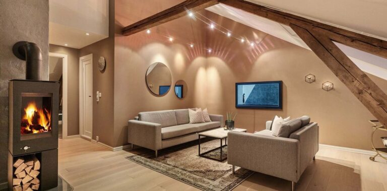 Stue med skråtak og synlige bærebjelker. Salongen med to sofaer mot hverandre er lys grå. Spotskikkenen i taket ir et vakkert lyssskinn på veggen