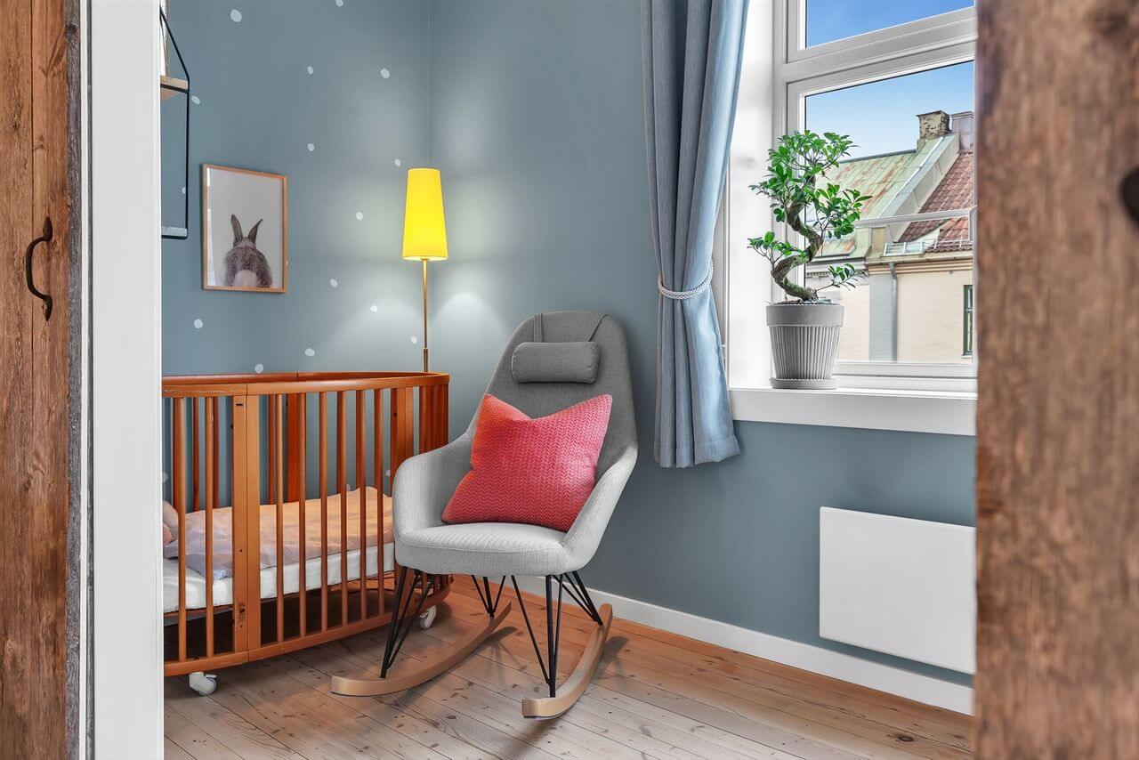 Babyseng i et soverom med blågrå veggfarge. Varm lys fra en gul lampe og ett bonsai tre i vinduskarmen gir ro og behag.