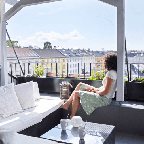 Stor sofa på takterrasse. Jente sitter å nyter sola og utsikten over byens tak